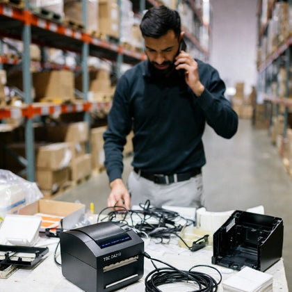 Man in a warehouse using a phone next to electronic equipment on a table.